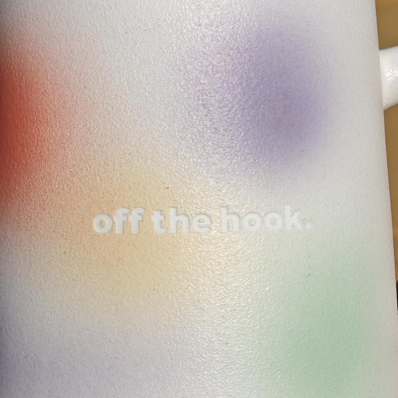 K&ouml;nnte beinhalten: Wei&szlig;er Becher mit einem farbenfrohen, abstrakten Design in Orange, Gelb, Lila und Gr&uuml;n. Der Schriftzug "off the hook." ist auf der Oberfl&auml;che des Bechers gepr&auml;gt. Der Becher hat einen Henkel und eine glatte, strukturierte Oberfl&auml;che.