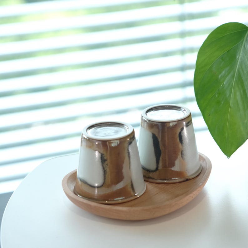 以下が含まれることがあります： 白ベースに茶色と黒の釉薬を施した2つのセラミックカップが、木製のトレイに置かれています。カップはユニークなデザインです。楕円形のトレイは明るい色の木材で作られています。