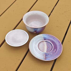 May include: A ceramic tea set with a cup, a small lid, and a saucer. The cup and lid are light pink with a white rim. The saucer is divided into white and purple sections with a pink rim. The set has a speckled glaze.
