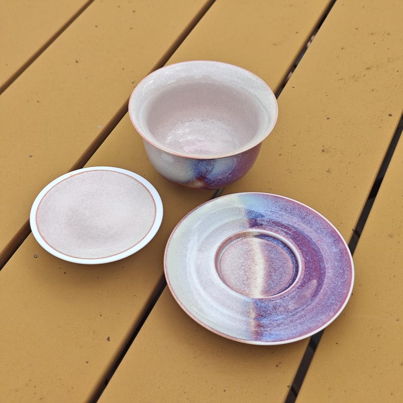 May include: A ceramic tea set with a cup, a small lid, and a saucer. The cup and lid are light pink with a white rim. The saucer is divided into white and purple sections with a pink rim. The set has a speckled glaze.
