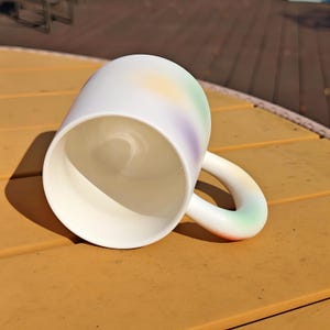 K&ouml;nnte beinhalten: Wei&szlig;e Keramiktasse mit rundem Henkel. Die Tasse hat einen Farbverlauf aus Pastellfarben, darunter Gelb, Gr&uuml;n und Lila. Die Tasse steht auf einer gelben Oberfl&auml;che.