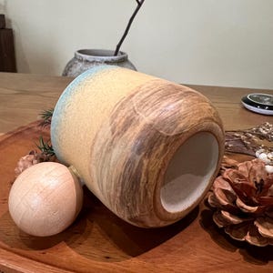 May include: A ceramic mug with a cylindrical shape, featuring a blend of tan, brown, and blue hues. The mug is placed on a wooden tray with a wooden ball and a pine cone. The mug's design has a textured, organic appearance.