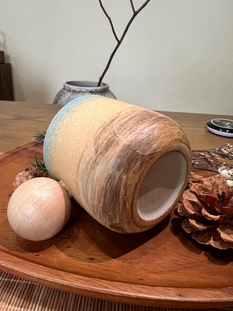 May include: A ceramic mug with a cylindrical shape, featuring a blend of tan, brown, and blue hues. The mug is placed on a wooden tray with a wooden ball and a pine cone. The mug's design has a textured, organic appearance.