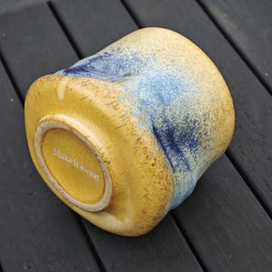 Peut inclure: Une tasse en c&eacute;ramique de forme unique, avec une base jaune et des accents bleus mouchet&eacute;s. Le fond de la tasse porte l'inscription "Make it sweet". La tasse est pos&eacute;e sur une surface en bois sombre.