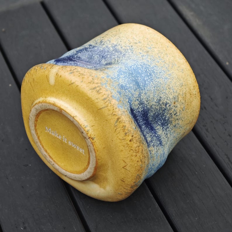 Peut inclure: Une tasse en c&eacute;ramique de forme unique, avec une base jaune et des accents bleus mouchet&eacute;s. Le fond de la tasse porte l'inscription "Make it sweet". La tasse est pos&eacute;e sur une surface en bois sombre.