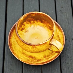 May include: A ceramic teacup and saucer set. The cup and saucer are a warm yellow color with brown trim. The cup has a handle and sits on a matching saucer. The set is placed on a dark wooden surface.
