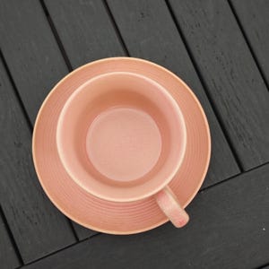 May include: Overhead view of a pink teacup and saucer set. The cup has a handle and sits on a matching saucer. Both pieces have a textured, ridged design. The set is a soft, muted pink color, perfect for serving tea or coffee.