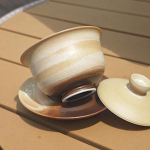 May include: A ceramic tea bowl with a lid and saucer, featuring a gradient of cream, beige, and dark brown tones. The bowl has a wide rim and a small footed base, while the matching saucer and lid display a consistent earthy color palette.