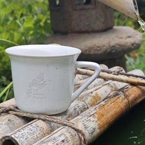 May include: A white ceramic mug with a handle, featuring a pagoda graphic and the text "Yu Garden Shanghai." The mug has dark speckles and rests on a bamboo structure. The background is blurred with green foliage.