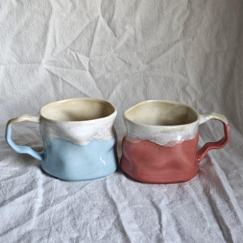 May include: Two handmade ceramic mugs. One is light blue, the other is a reddish-pink. Both have a white and beige rim and handle. The mugs have a textured, organic shape, suggesting a rustic or artisanal style.