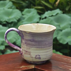 May include: A handmade ceramic mug with a purple and cream swirl pattern. The mug has a rounded body, a curved handle, and a slightly flared rim. The mug is approximately 4 inches tall and 3.5 inches in diameter.