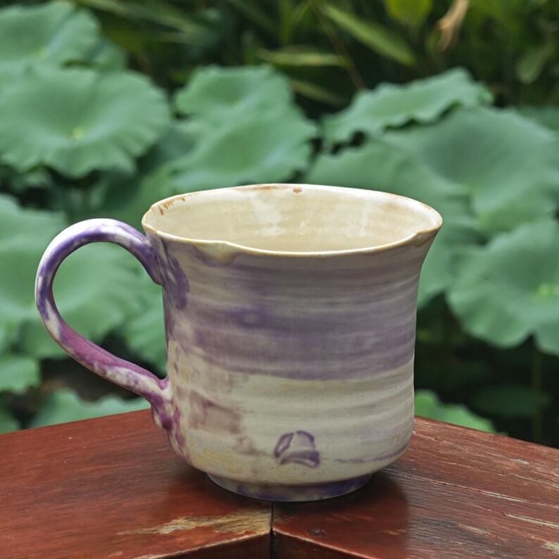 May include: A handmade ceramic mug with a purple and cream swirl pattern. The mug has a rounded body, a curved handle, and a slightly flared rim. The mug is approximately 4 inches tall and 3.5 inches in diameter.