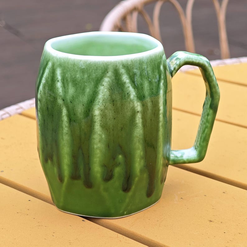 May include: A green ceramic mug with a textured surface and a square handle. The mug has a glossy finish with darker green drips, and it sits on a yellow surface. The mug is designed for hot or cold beverages.