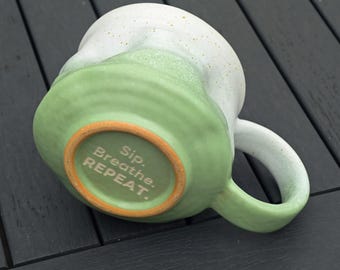 May include: A ceramic mug with a white top and a green base. The mug has a rounded handle and the words "Sip. Breathe. REPEAT." are printed on the bottom in a light color. The mug is resting on a dark gray surface.