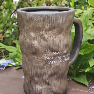 Puede incluir: Una taza de cer&aacute;mica alta y cil&iacute;ndrica con un exterior de color marr&oacute;n oscuro texturizado que se asemeja a la corteza de un &aacute;rbol. La taza tiene un asa en el lado derecho y un texto impreso en blanco que dice: Michael- Morning Person (After Coffee).