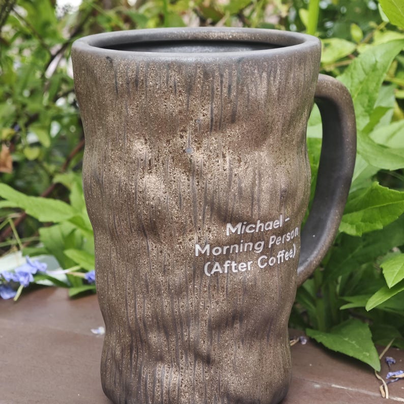 Puede incluir: Una taza de cer&aacute;mica alta y cil&iacute;ndrica con un exterior de color marr&oacute;n oscuro texturizado que se asemeja a la corteza de un &aacute;rbol. La taza tiene un asa en el lado derecho y un texto impreso en blanco que dice: Michael- Morning Person (After Coffee).