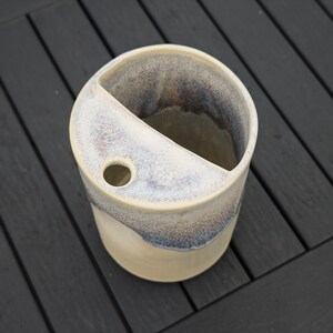 May include: A cylindrical ceramic vessel featuring a cream-colored base and a mottled blue and white glazed top section. The top surface is partially covered by a flat ledge with a single circular opening, creating a distinct divided interior space.