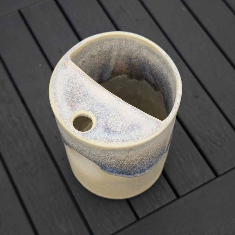 May include: A cylindrical ceramic vessel featuring a cream-colored base and a mottled blue and white glazed top section. The top surface is partially covered by a flat ledge with a single circular opening, creating a distinct divided interior space.