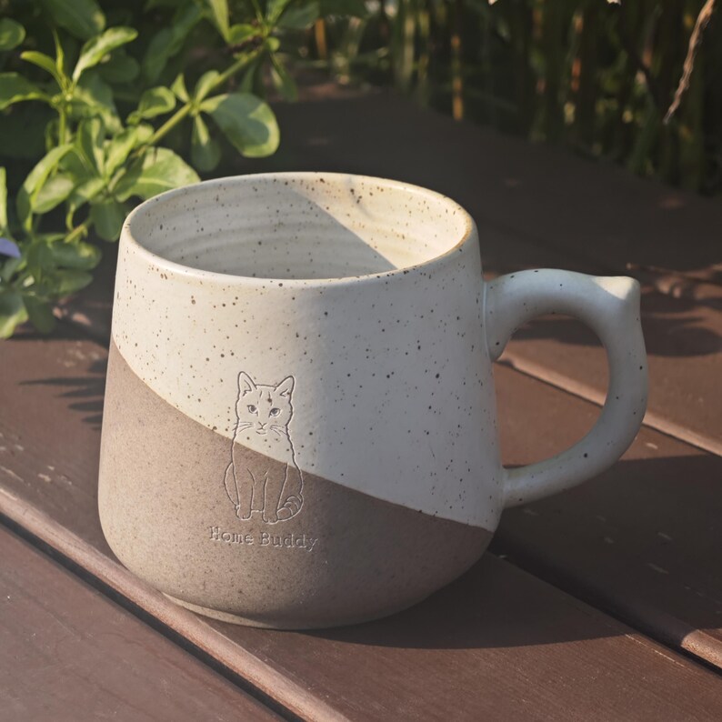 May include: A two-toned ceramic mug featuring a speckled off-white upper section and a solid light brown lower section. An engraved illustration of a seated cat is centred on the front, accompanied by the text Home Buddy printed below the cat.