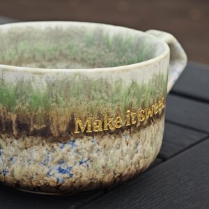 May include: A ceramic mug with a textured, earthy design in shades of green, brown, and blue. The mug features the gold text "Make it sweet" on its side. The mug has a handle and a rustic aesthetic.