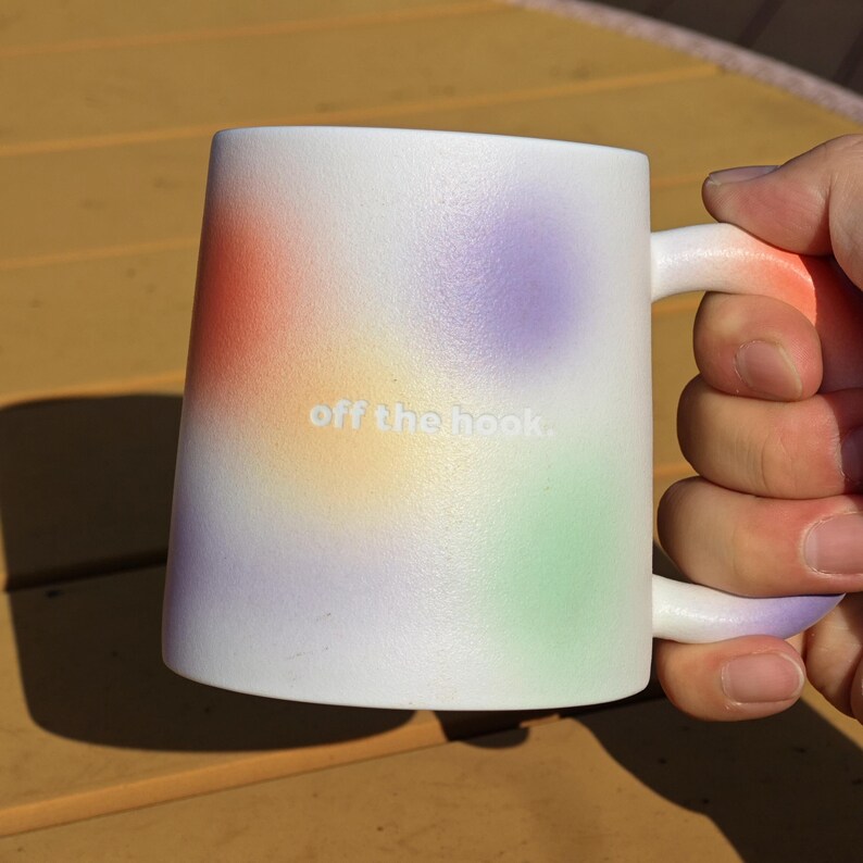 K&ouml;nnte beinhalten: Wei&szlig;er Keramikbecher mit einem farbenfrohen Farbverlauf in Orange, Gelb, Lila und Gr&uuml;n. Der Becher tr&auml;gt den Text "off the hook". Der Griff und der Rand des Bechers sind wei&szlig;.