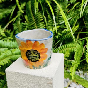 May include: A small, handmade ceramic cup with a sunflower design. The cup has a light gray base, a blue rim, and a vibrant orange and yellow sunflower painted on the front. Green leaves and a black center complete the floral design.