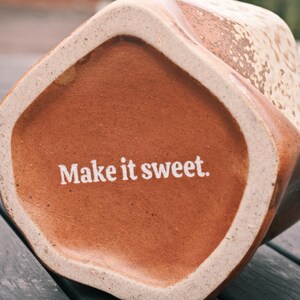 Peut inclure: Gros plan sur le fond d'une tasse en c&eacute;ramique marron avec l'inscription "Make it sweet" imprim&eacute;e en blanc. La tasse a une forme g&eacute;om&eacute;trique et une surface textur&eacute;e, avec un bord de couleur claire.