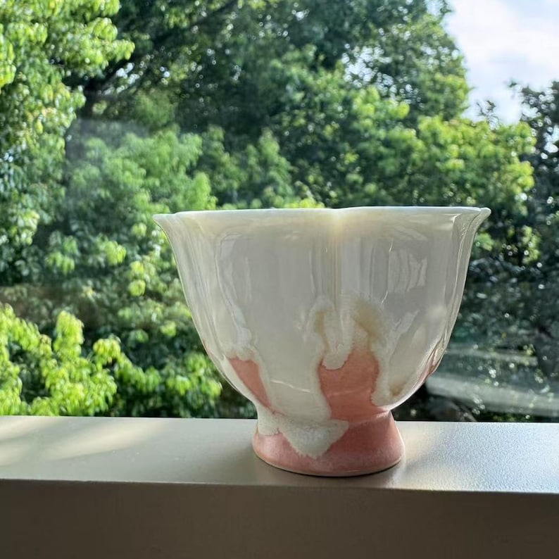 Pode incluir: Uma tigela de cer&acirc;mica branca com uma borda alargada e uma base rosa. A tigela tem uma forma &uacute;nica e um acabamento brilhante. O interior e o exterior da tigela t&ecirc;m um esmalte branco com detalhes rosa e bege. A tigela est&aacute; sobre uma superf&iacute;cie cinza.