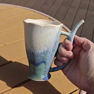 May include: A handmade ceramic mug with a unique handle design. The mug features a wavy rim and a gradient of colors, transitioning from white to blue and green. The mug is held in a hand, showcasing its size and shape.