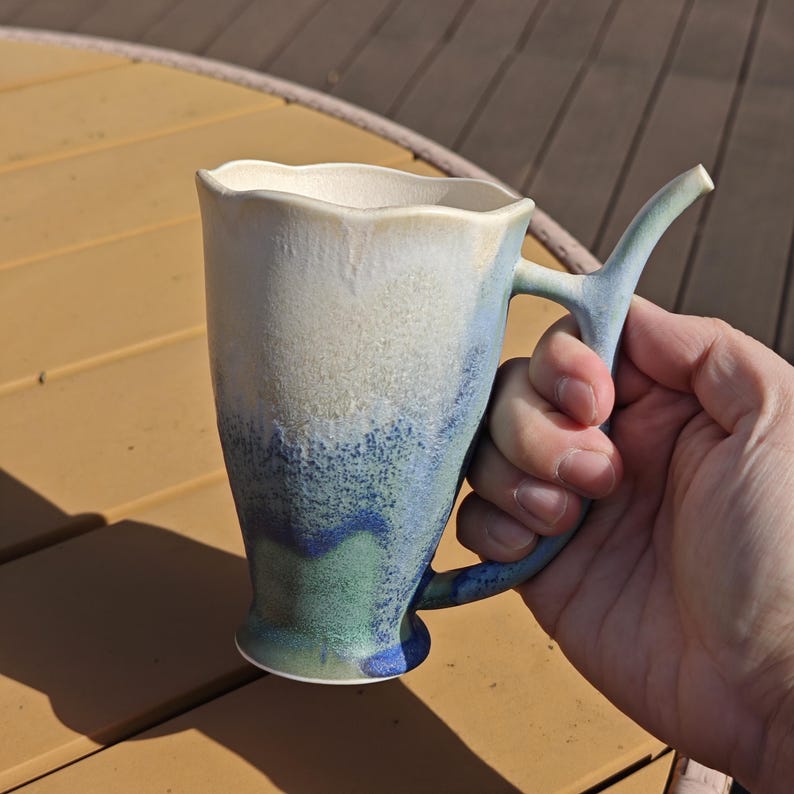 May include: A handmade ceramic mug with a unique handle design. The mug features a wavy rim and a gradient of colors, transitioning from white to blue and green. The mug is held in a hand, showcasing its size and shape.