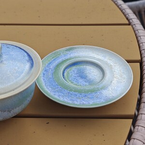 May include: A ceramic plate and a small bowl with a lid. The plate has concentric circles of blue, green, and white. The bowl and lid are light blue with a white rim. The items are on a brown surface.