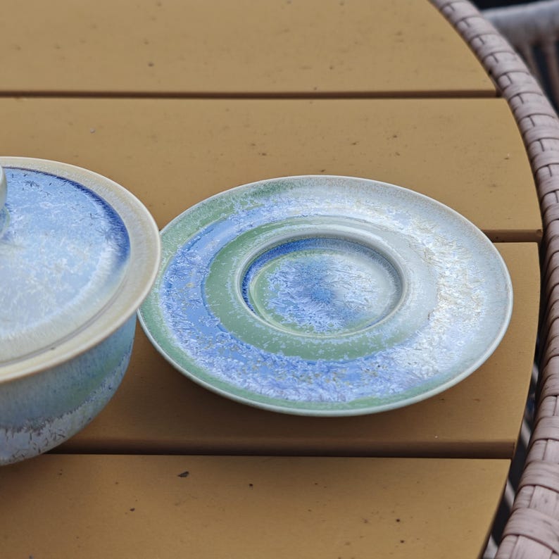 May include: A ceramic plate and a small bowl with a lid. The plate has concentric circles of blue, green, and white. The bowl and lid are light blue with a white rim. The items are on a brown surface.