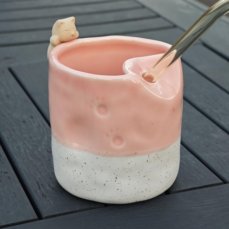 May include: A pink and white ceramic mug with a cat figurine on the rim. The mug has paw print indentations and a unique spout. The bottom half is white with black speckles. A silver metal object is pouring liquid into the mug.