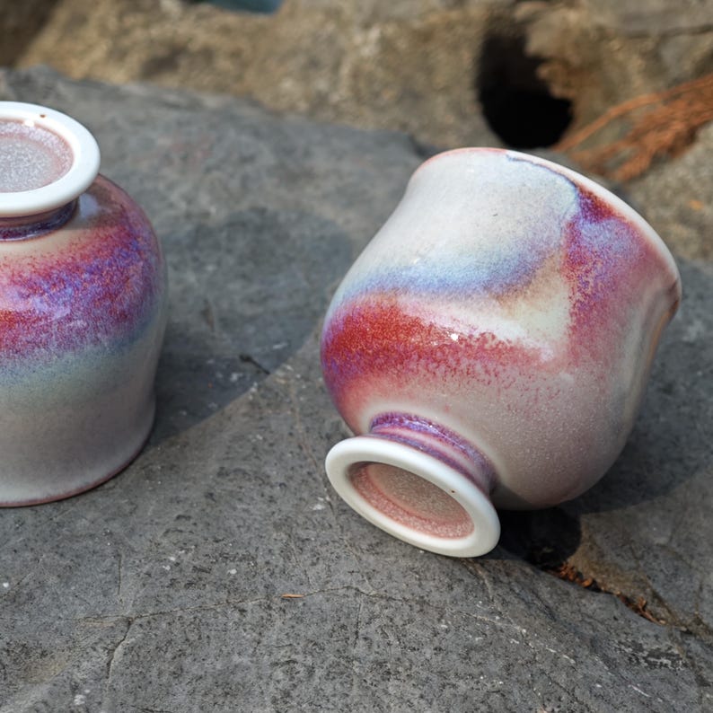May include: Two ceramic teacups with a unique glaze. The cups have a rounded shape with a white rim and base. The glaze features a blend of red, purple, and blue hues, creating a marbled effect. The cups are placed on a gray stone surface.