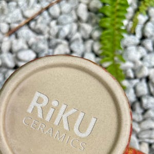 Op de afbeelding: Close-up van de bodem van een keramische mok met de in reli&euml;f aangebrachte tekst "Riku Ceramics" in het wit. De basis van de mok is lichtbeige van kleur en de rand heeft een rood en oranje ontwerp. De achtergrond heeft witte kiezels en groen gebladerte.