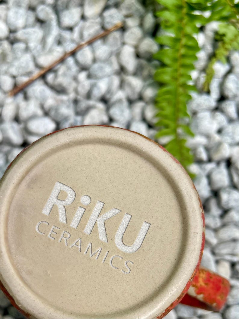 Op de afbeelding: Close-up van de bodem van een keramische mok met de in reli&euml;f aangebrachte tekst "Riku Ceramics" in het wit. De basis van de mok is lichtbeige van kleur en de rand heeft een rood en oranje ontwerp. De achtergrond heeft witte kiezels en groen gebladerte.