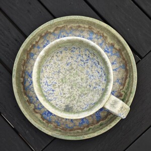 May include: Overhead view of a ceramic cup and saucer set. The cup and saucer have a speckled pattern in shades of green, blue, and brown. The cup has a handle and sits on a matching saucer. The set is placed on a dark wooden surface.