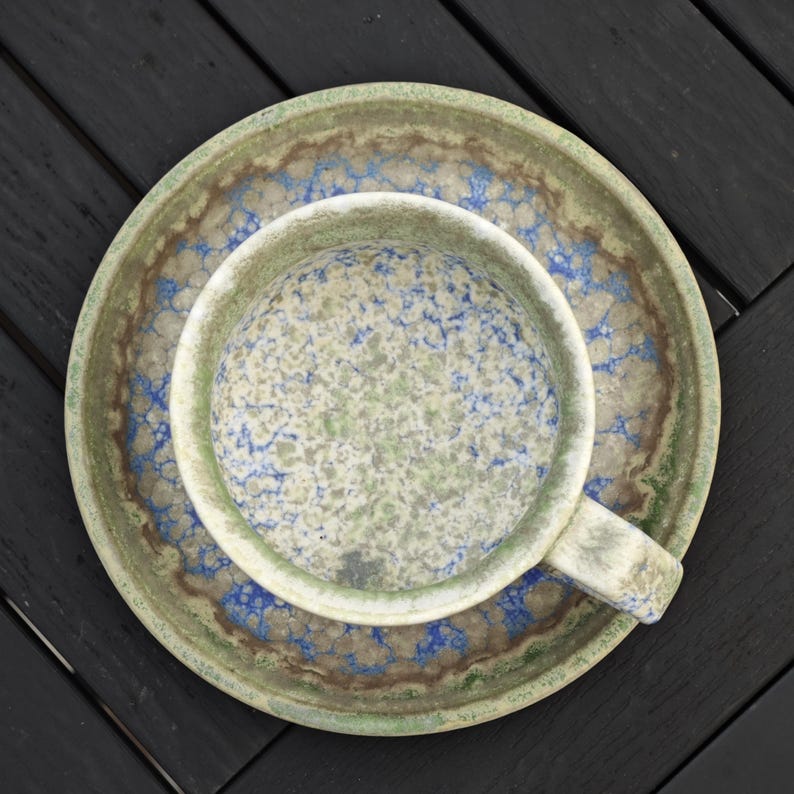 May include: Overhead view of a ceramic cup and saucer set. The cup and saucer have a speckled pattern in shades of green, blue, and brown. The cup has a handle and sits on a matching saucer. The set is placed on a dark wooden surface.