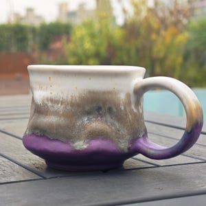 Op de afbeelding: Een handgemaakte keramische mok met een unieke glazuur. De mok heeft een witte bovenkant, overgaand in bruin- en grijstinten, en eindigt met een levendige paarse basis. Het handvat heeft dezelfde kleur. Perfect voor koffie of thee.