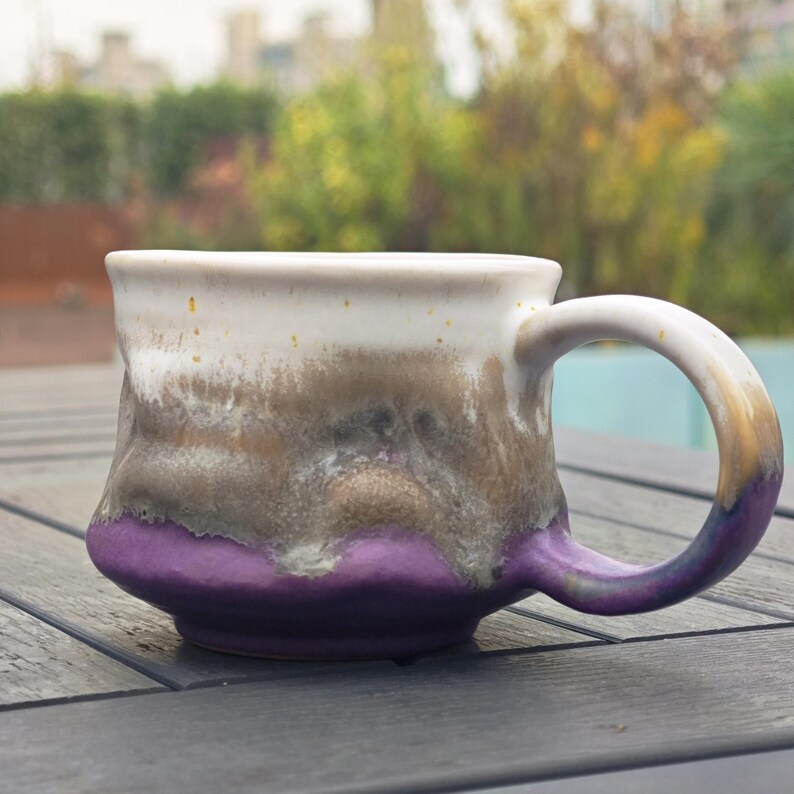 Op de afbeelding: Een handgemaakte keramische mok met een unieke glazuur. De mok heeft een witte bovenkant, overgaand in bruin- en grijstinten, en eindigt met een levendige paarse basis. Het handvat heeft dezelfde kleur. Perfect voor koffie of thee.