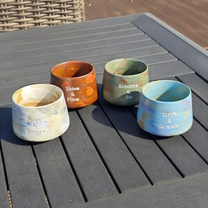 May include: Four ceramic cups with inspirational phrases. The cups are in shades of blue, green, orange, and cream. Each cup has a different phrase: "Rise & Focus", "Shine & Flow", "Breathe & Be", and "Drift & Dream".