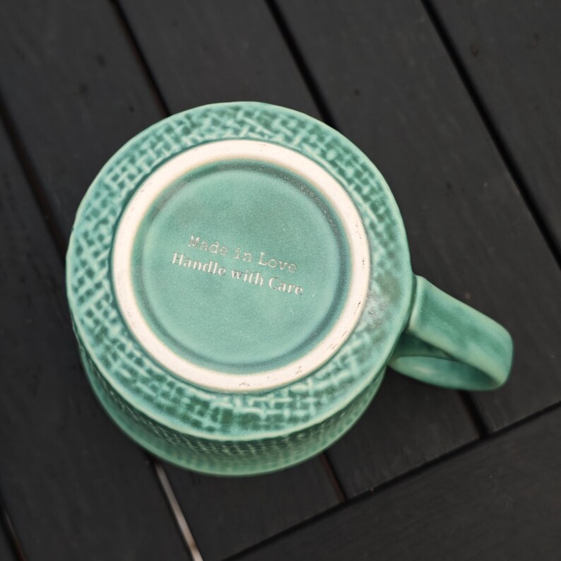 May include: A teal ceramic mug with a textured exterior and a handle. The bottom of the mug features the text "Made in Love Handle with Care". The mug is set against a dark wooden surface.
