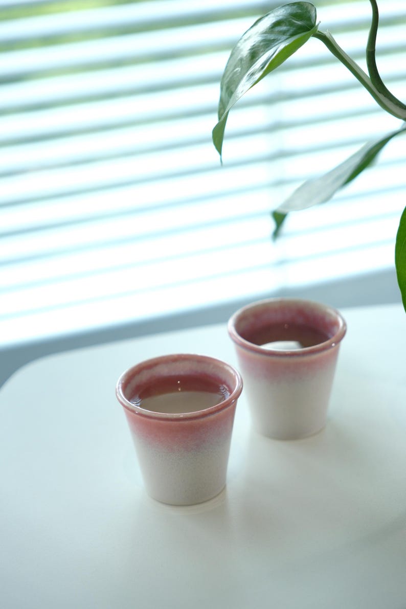 May include: Two small ceramic cups with a gradient pink and white design. The cups are filled with a clear liquid. The cups are on a white surface, with a plant in the background.