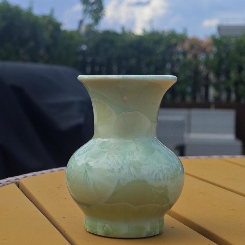 Puede incluir: Un jarr&oacute;n de cer&aacute;mica verde p&aacute;lido con cuerpo bulboso y borde acampanado. El jarr&oacute;n tiene un acabado brillante con sutiles patrones cristalinos, verde claro y blanco. El jarr&oacute;n mide unos 15 cm de alto.