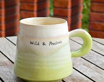 May include: A ceramic mug with a rounded base and a large, curved handle. The mug transitions from a creamy white at the top to a light green at the bottom, with the words "Wild & Precious" in black script. The mug is on a wooden surface.
