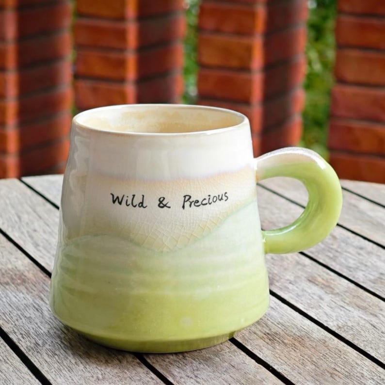 May include: A ceramic mug with a rounded base and a large, curved handle. The mug transitions from a creamy white at the top to a light green at the bottom, with the words "Wild & Precious" in black script. The mug is on a wooden surface.