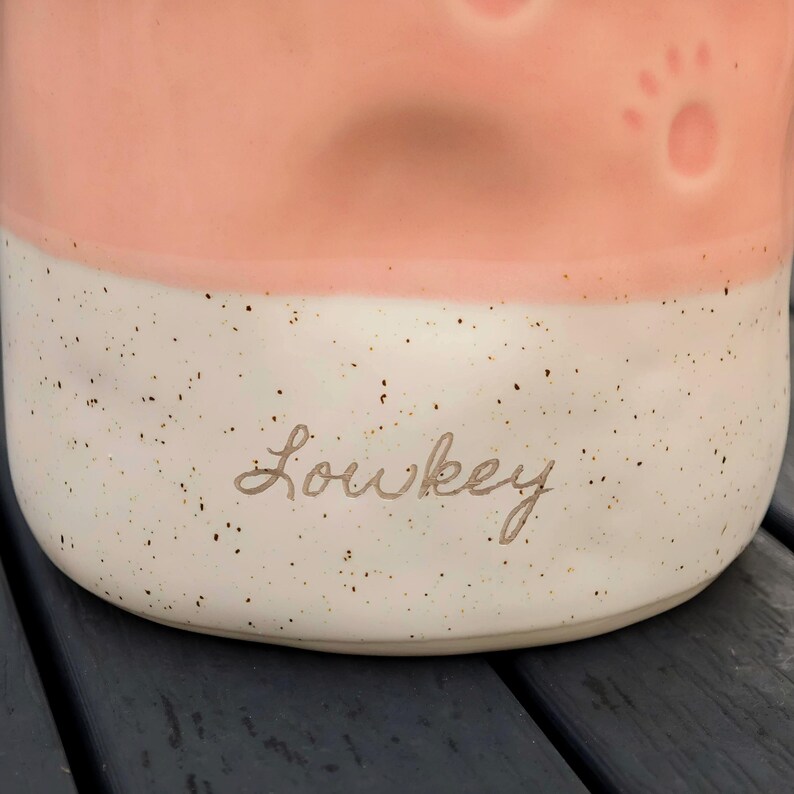 May include: A ceramic mug with a two-tone design. The top half is a soft pink, and the bottom half is speckled white. The word "Lowkey" is written in cursive on the bottom. Paw prints are embossed on the pink section.