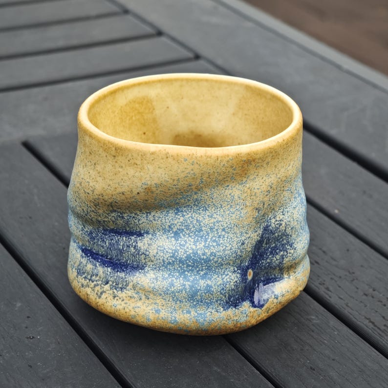 Peut inclure: Une tasse en c&eacute;ramique faite &agrave; la main avec une surface textur&eacute;e. La tasse pr&eacute;sente un m&eacute;lange de couleurs, notamment un bord beige clair, une section m&eacute;diane bleut&eacute;e et un aspect mouchet&eacute;. La tasse est de forme l&eacute;g&egrave;rement irr&eacute;guli&egrave;re.