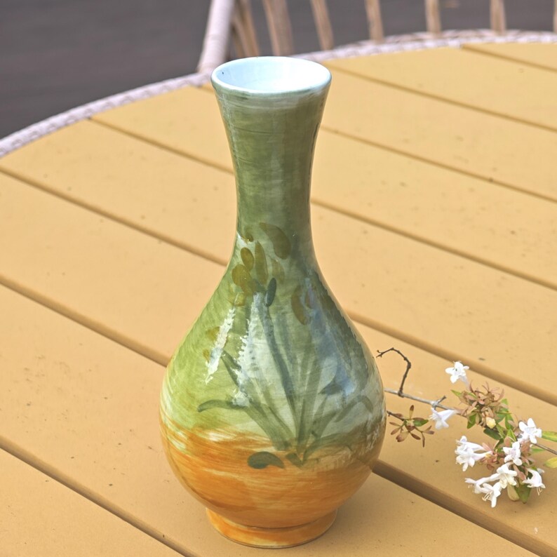 K&ouml;nnte beinhalten: Eine handbemalte Keramikvase mit schlankem Hals und bauchigem K&ouml;rper. Die Vase hat einen Farbverlauf, der von einem warmen orangefarbenen Sockel zu einem gr&uuml;nen oberen Bereich mit floralen Akzenten &uuml;bergeht. Der Innenraum ist hellblau.