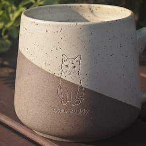 May include: A two-toned ceramic mug featuring a speckled cream upper section and a solid brown lower section. An embossed illustration of a seated cat is centred on the front, positioned above the engraved text Home Buddy in a simple font.